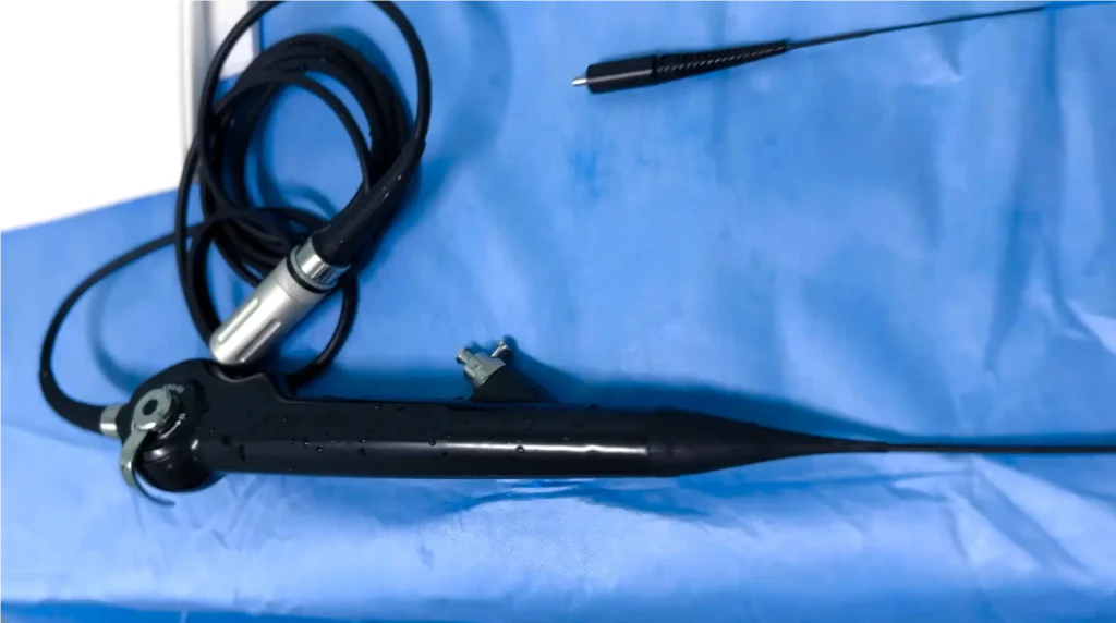 Cistoscópio flexível digital preto, com cabo e conector, posicionado sobre um campo cirúrgico azul. O equipamento está com gotas d’água, indicando recente higienização ou uso.
