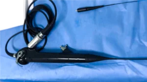 Cistoscópio flexível digital preto, com cabo e conector, posicionado sobre um campo cirúrgico azul. O equipamento está com gotas d’água, indicando recente higienização ou uso.